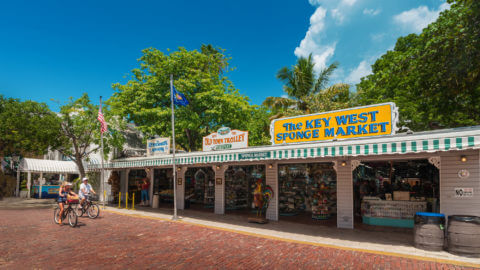 Two people biking along Mallory Square in front of various shops in Key West, FL