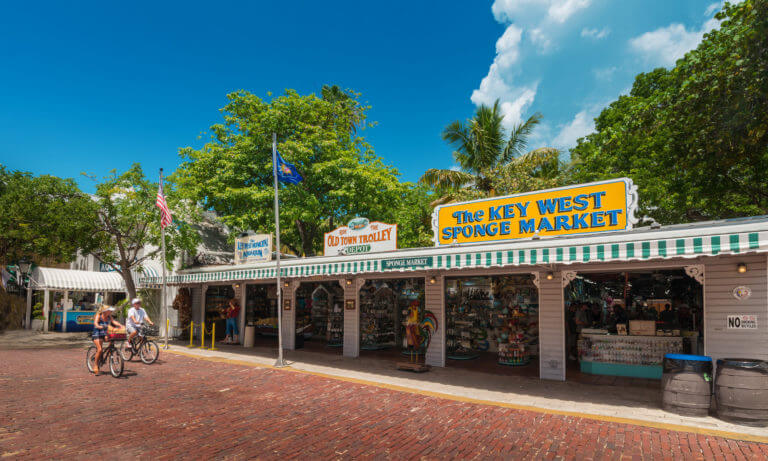 Two people biking along Mallory Square in front of various shops in Key West, FL