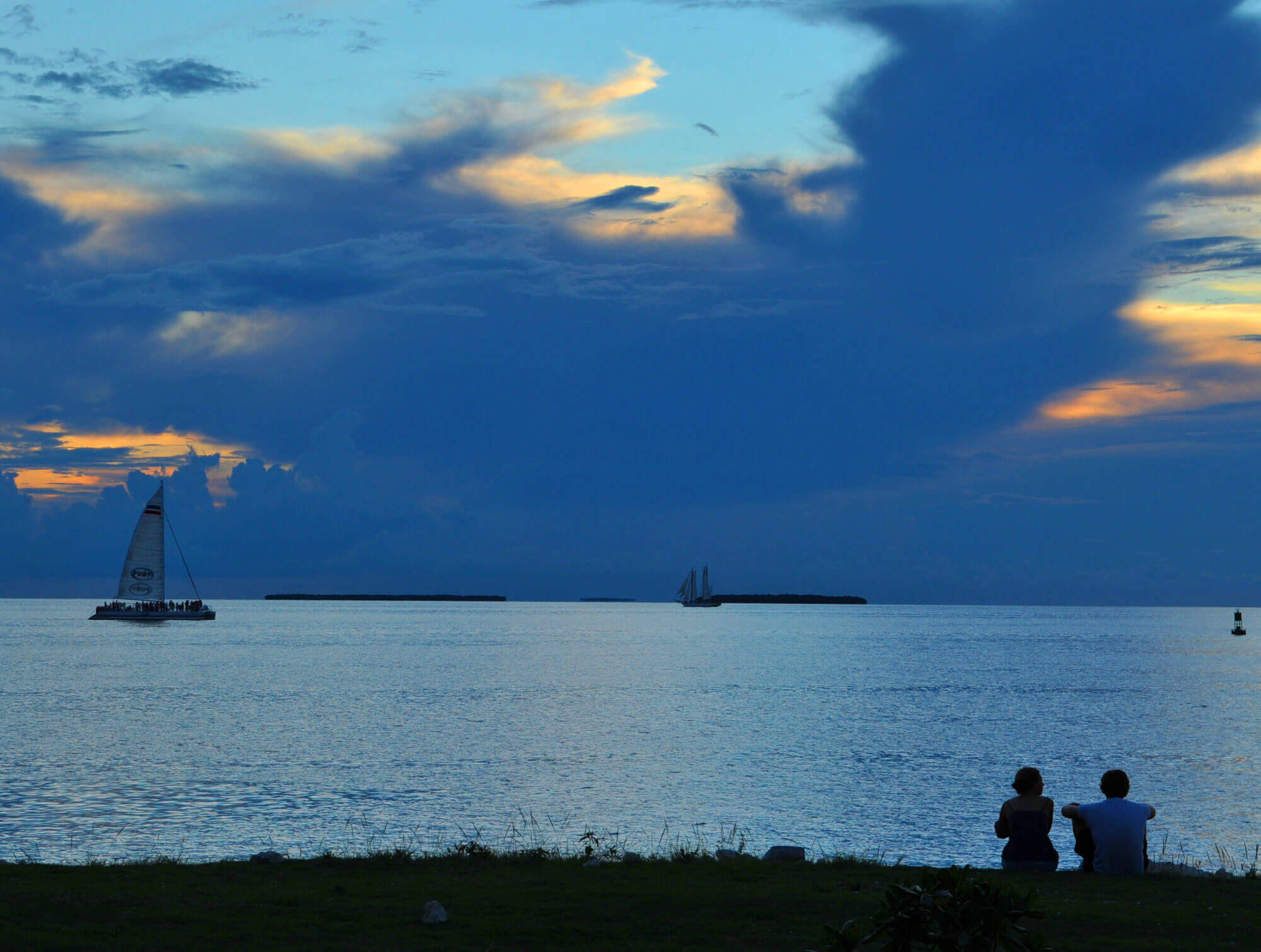 Two people sitting down at the ocean's edge in Key West, FL looking at a Fury catamaran sailing by