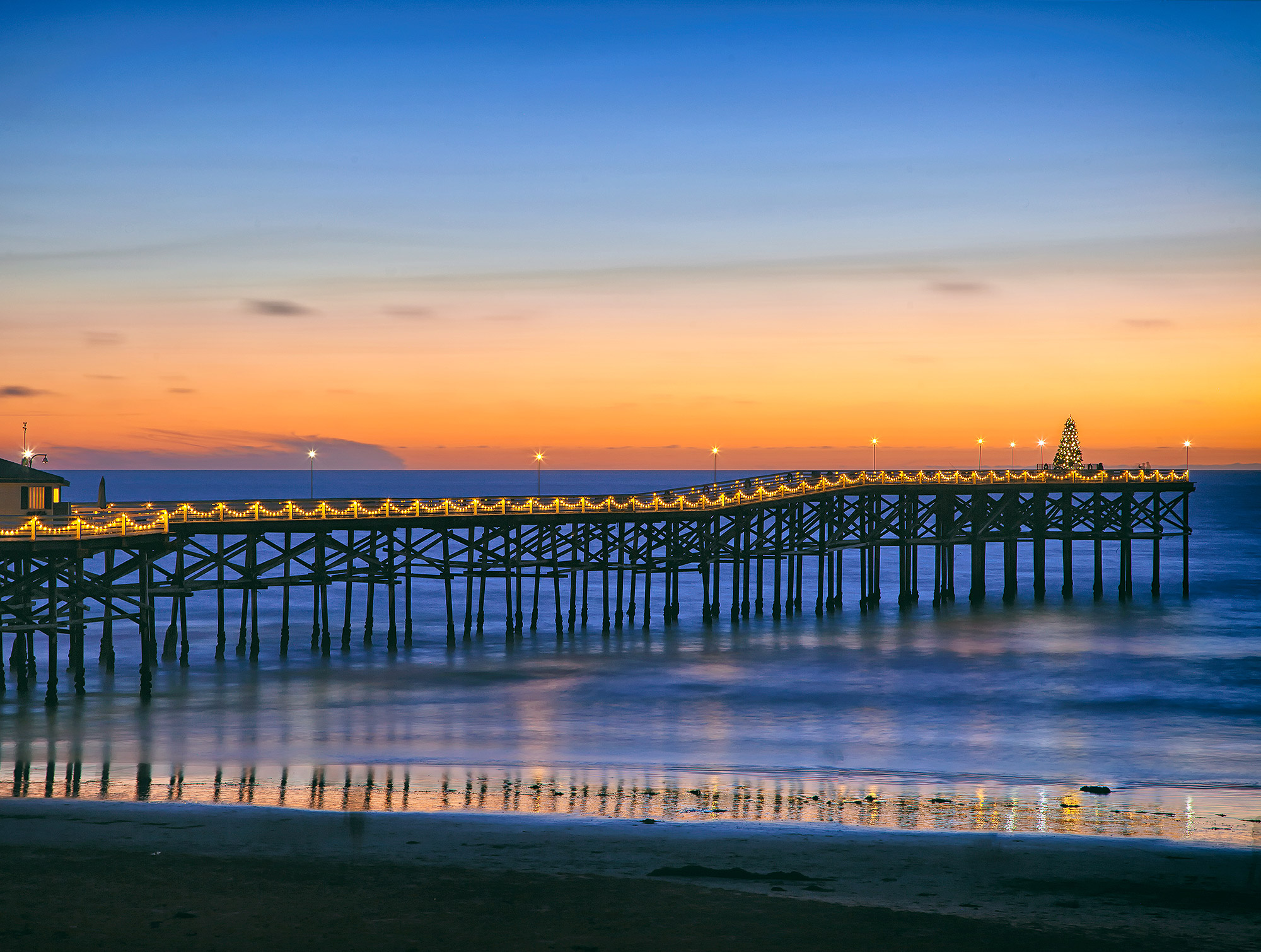 San Diego Crystal Pier at sunset featuring holiday lights strung on pier, the ocean and a Christmas tree at the end of pier