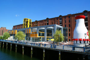picture of front of Boston Children's Museum building made up of brick and glass, Boston Harbor in the foreground, people sitting on picnic tables in front of museum and a giant structure on the right shaped like a milk bottle with windows and awnings
