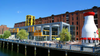 picture of front of Boston Children's Museum building made up of brick and glass, Boston Harbor in the foreground, people sitting on picnic tables in front of museum and a giant structure on the right shaped like a milk bottle with windows and awnings