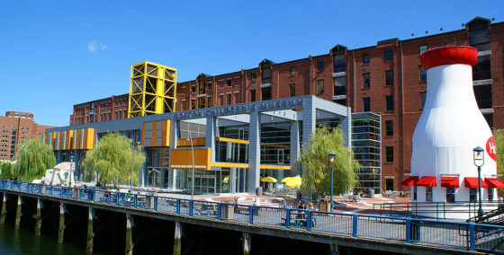 picture of front of Boston Children's Museum building made up of brick and glass, Boston Harbor in the foreground, people sitting on picnic tables in front of museum and a giant structure on the right shaped like a milk bottle with windows and awnings