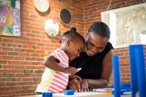 mother and daughter at Boston Children's Museum working on an art project at a desk with markers and blocks and a brick wall in the background with pieces of art on wall