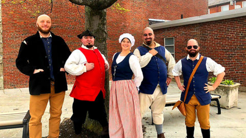 a group of one woman and five men in Boston standing in front of a tree and wearing colonial period clothing