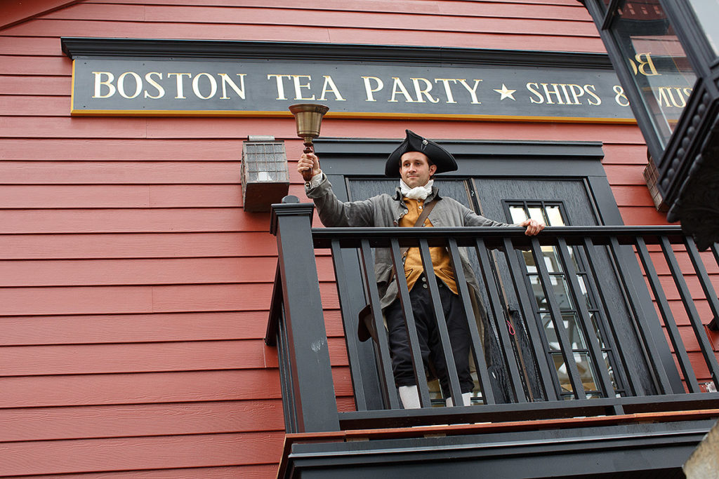 Boston actor dressed in colonial clothes and standing on a balcony ringing a bell; behind him is a sign that reads 'Boston Tea Party Ships & Museum'