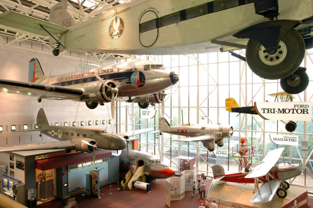 exhibit of various types of air planes; some of them suspended by ceiling and others sitting on the ground with a glass wall background