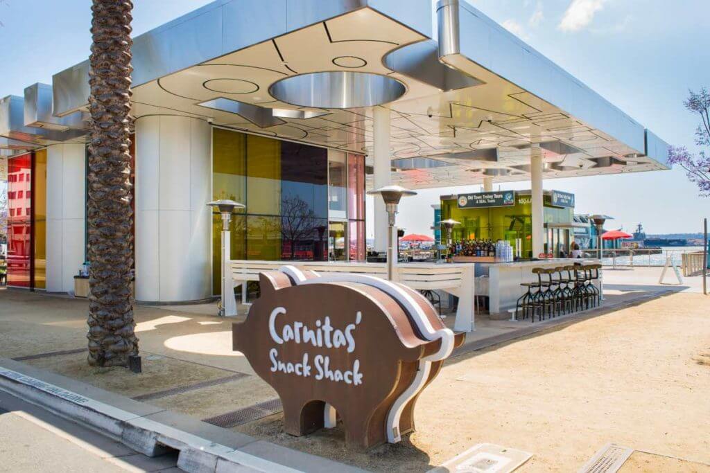 silhouette sign of a pig in the foreground with the words 'Carnitas' Snack Shack', open air restaurant with bar and chairs, and building ocean in the background