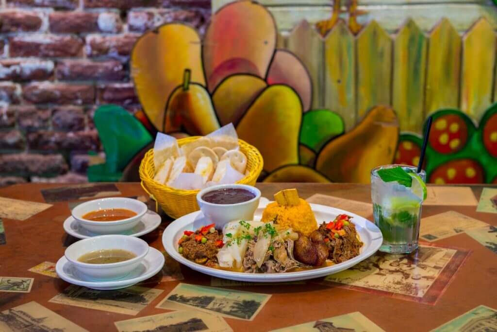 picture of a table at El Meson de Pepe showing bread basket, soups, drink and a dish consisting of various meats, rice, beans and plantains; table is decorated with pictures and background has painted artwork showing a picket fence and tropical fruit