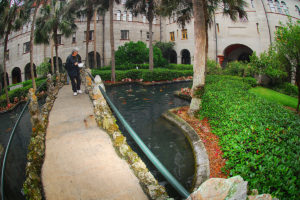 courtyard at Lightner Museum featuring a lady on a footbridge feeding koi fish swimming in pond and surrounded by foliage