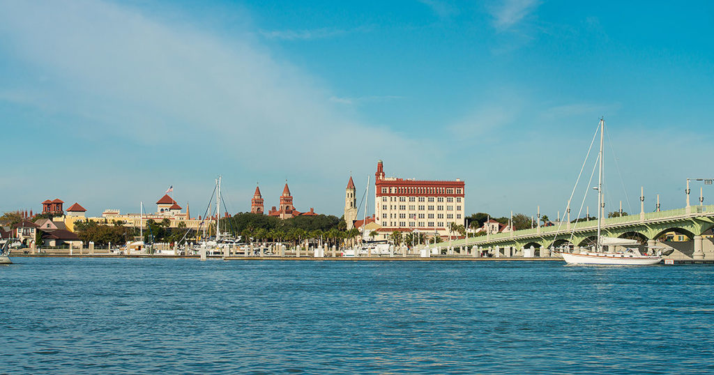 daytime photo of St. Augustine skyline featuring several ancient looking buildings and towers, a sailboat and bridge of lions to the right, and ocean on the foreground