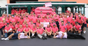 a group of cast members all dressed in pink shirts and standing in front of a San Diego trolley and a large inflatable chair that reads 'American Cancer Society Making Strides Against Breast Cancer'
