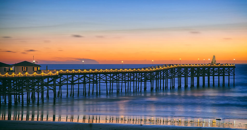 San Diego Crystal Pier at sunset featuring holiday lights strung on pier, the ocean and a Christmas tree at the end of pier