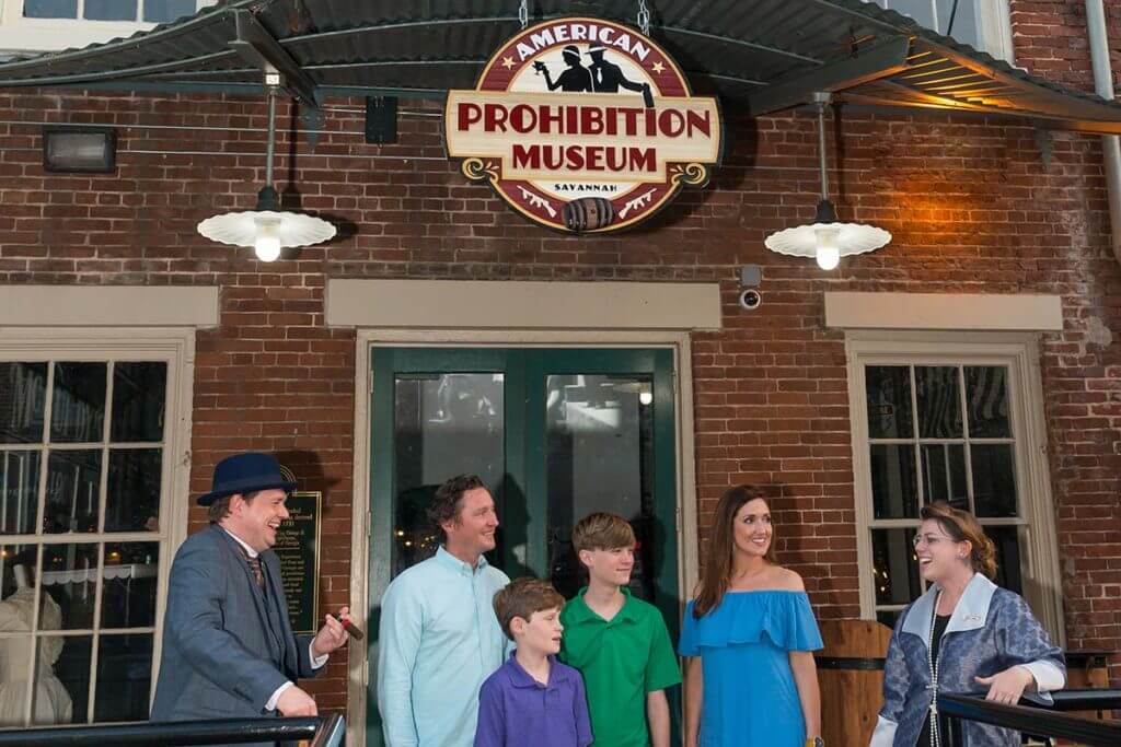 A smiling family of four standing in front of the American Prohibition Museum in Savannah flanked by a man and woman in Prohibition era clothing