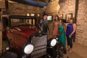 Image of a maroon colored vintage 1920s era automobile next to three guests and a man in a period accurate blue 3-piece suit laughing with a cigar in his hand in the American Prohibition Museum in Savannah,GA
