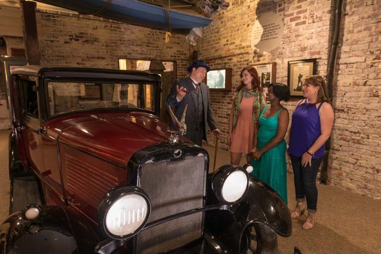 Image of a maroon colored vintage 1920s era automobile next to three guests and a man in a period accurate blue 3-piece suit laughing with a cigar in his hand in the American Prohibition Museum in Savannah,GA