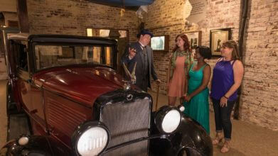 Image of a maroon colored vintage 1920s era automobile next to three guests and a man in a period accurate blue 3-piece suit laughing with a cigar in his hand in the American Prohibition Museum in Savannah,GA