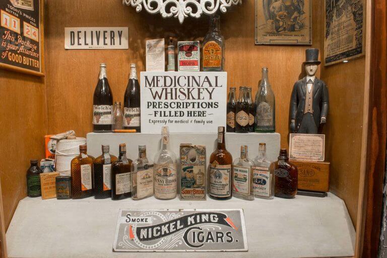 A display of antique liquor bottles next to a sign that reads 'Medicinal Whiskey prescriptions filled here' at the American Prohibition Museum in Savannah, GA