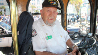 An Old Town Trolley conductor named Bill Fox sitting in the trolley's drivers chair smiling and wearing a hat that says 'Korea-Vietnam Veteran'