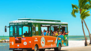 Riders inside an Old Town Trolley waving as they drive down a beach in Key West