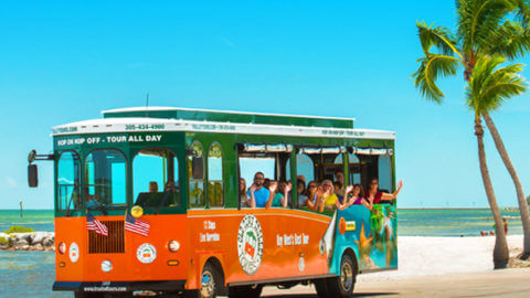Riders inside an Old Town Trolley waving as they drive down a beach in Key West