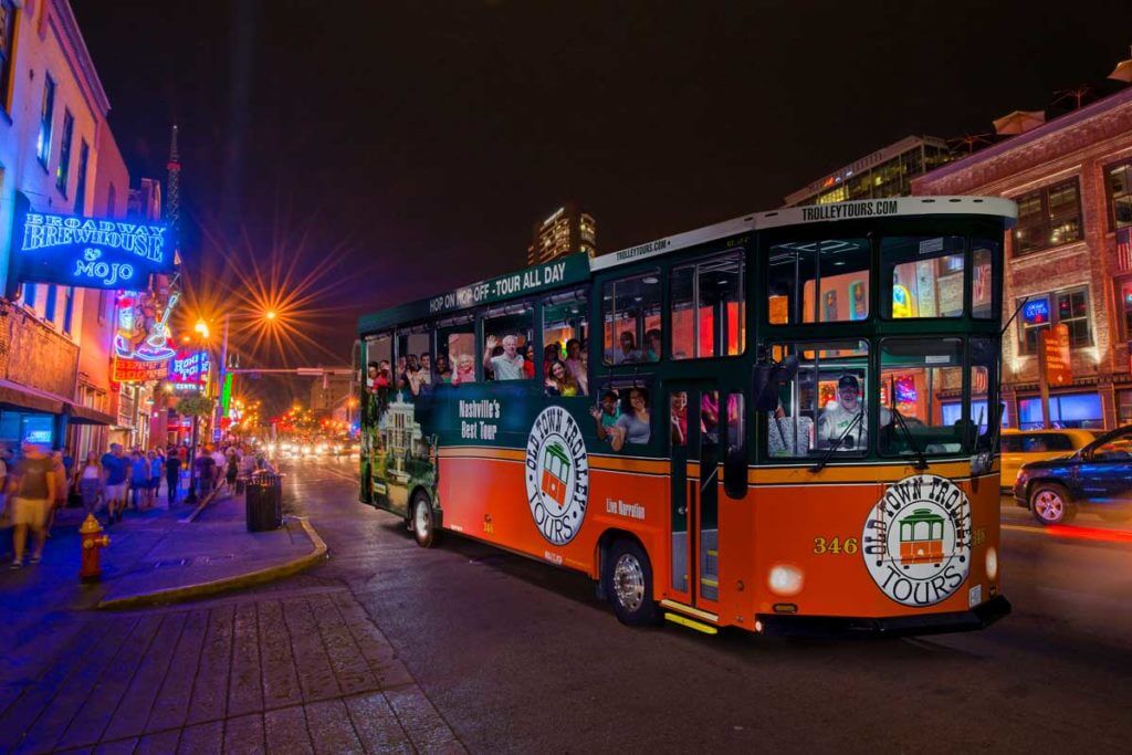 Night time picture of Old Town Trolley packed with people waving and smiling down Lower Broadway in Nashville, TN
