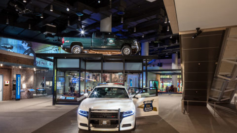 national law enforcement museum exhibit featuring a police car in the center with a door open and the words 'Indiana State Police' and in the background, glass case with a truck mounted on the top