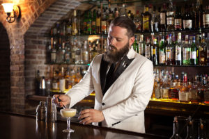 picture showing a bartender in suit and tie fixing an alcoholic beverage with rows on liquor bottles on glass shelves on display behind him