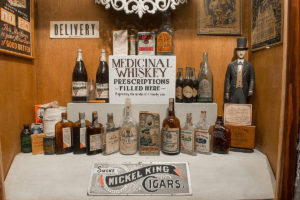 museum showing two rows of whiskey bottles, several art pieces on the wall, a sign that reads 'delivery', a sign that reads 'smoke nickel king cigars', and a sign that reads 'medicinal whiskey prescriptions filled here, expressly for medical & family use'
