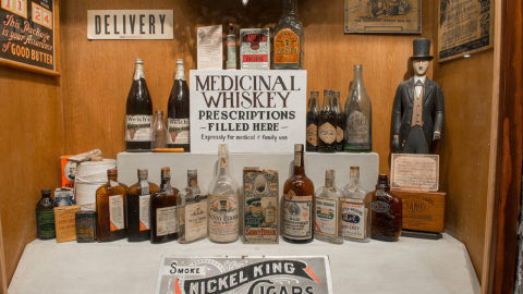 museum showing two rows of whiskey bottles, several art pieces on the wall, a sign that reads 'delivery', a sign that reads 'smoke nickel king cigars', and a sign that reads 'medicinal whiskey prescriptions filled here, expressly for medical & family use'