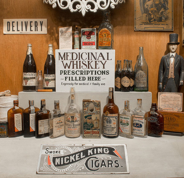 museum showing two rows of whiskey bottles, several art pieces on the wall, a sign that reads 'delivery', a sign that reads 'smoke nickel king cigars', and a sign that reads 'medicinal whiskey prescriptions filled here, expressly for medical & family use'