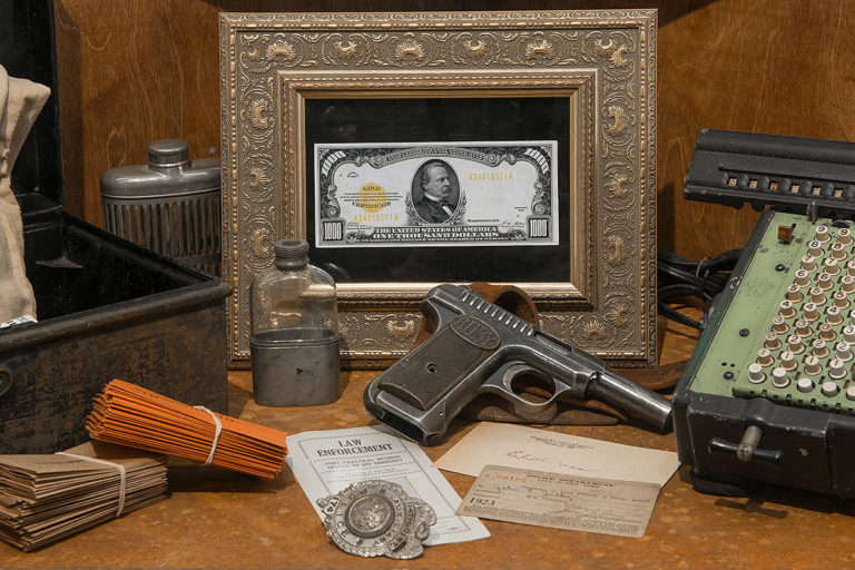 museum display showing a framed $1,000 bill, a gun, two old flask bottles, a counter, a tin box, notes, law enforcement badge.
