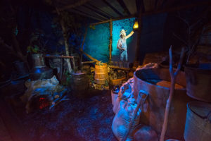 museum display showing a moonshiner camp at night with barrels, bottles and a fireplace and a hologram of a moonshiner in the background