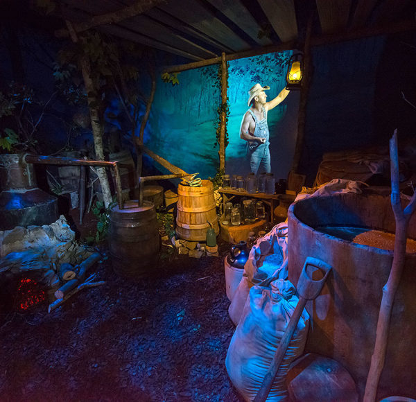 museum display showing a moonshiner camp at night with barrels, bottles and a fireplace and a hologram of a moonshiner in the background