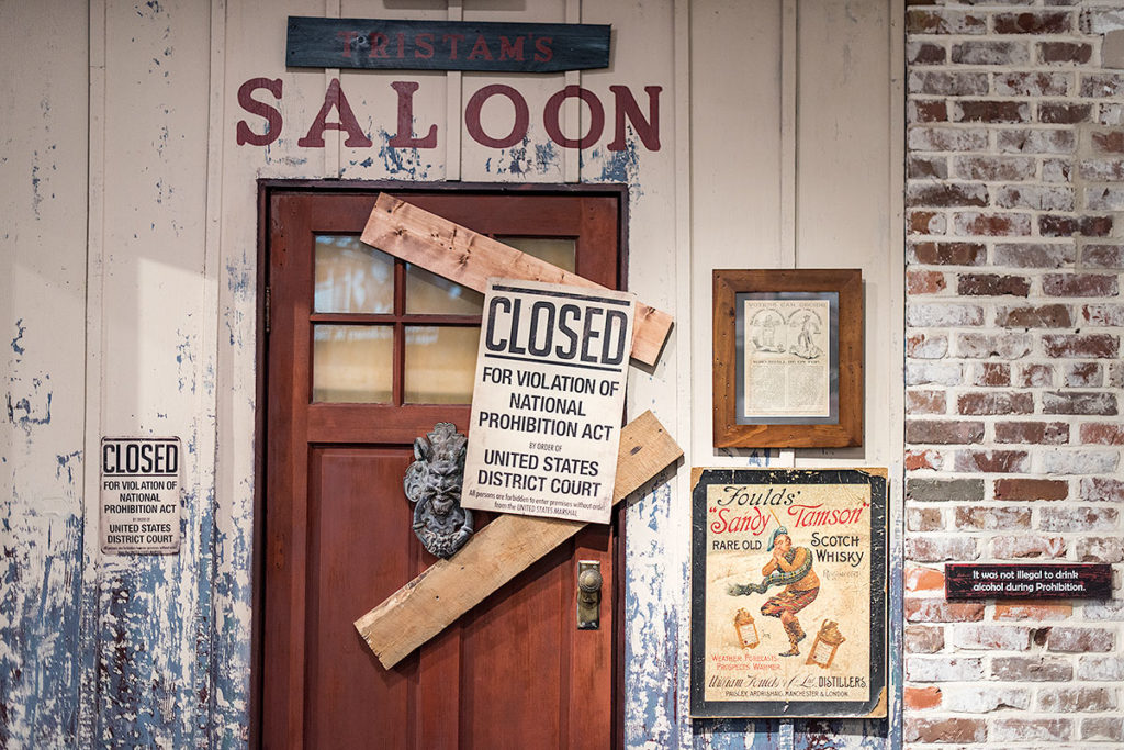 picture showing wood and brick wall, a door with the word 'saloon' above it, two boards blocking door and a sign that reads 'closed for violation of national prohibition act by order of United States District Court'