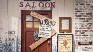 picture showing wood and brick wall, a door with the word 'saloon' above it, two boards blocking door and a sign that reads 'closed for violation of national prohibition act by order of United States District Court'