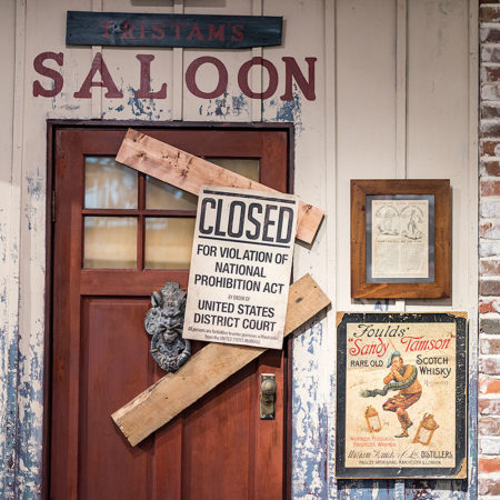 picture showing wood and brick wall, a door with the word 'saloon' above it, two boards blocking door and a sign that reads 'closed for violation of national prohibition act by order of United States District Court'
