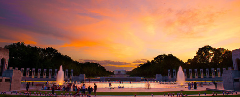 Sunset picture of WWII memorial in Washington DC made up of columns and fountains and the Lincoln Memorial far off in the background