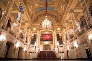 picture of boch center lobby featuring two floors, a set of carpeted stars, ornate walls and ceiling, a hanging crystal chandelier, and columns on second floor