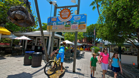 guests walking through Key West's Mallory Square made up of food booths and carts. In the background, there's a statue of a man riding on a bicycle, two barrels, a sign that says 'Mallory Square' and tables and chairs