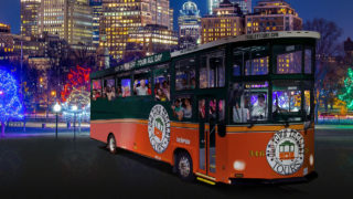 night time picture of trolley in front of Boston skyline and holiday lights