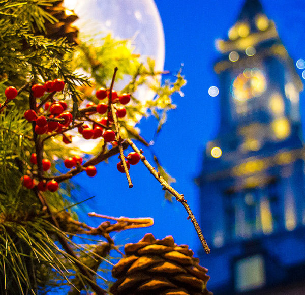 Nighttime Boston picture of holiday decorations including holly leaves and pinecones and a full moon and Faneuil Hall in the background