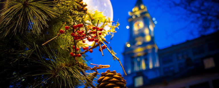 Nighttime Boston picture of holiday decorations including holly leaves and pinecones and a full moon and Faneuil Hall in the background