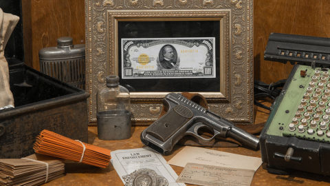 museum display showing a framed $1,000 bill, a gun, two old flask bottles, a counter, a tin box, notes, law enforcement badge.