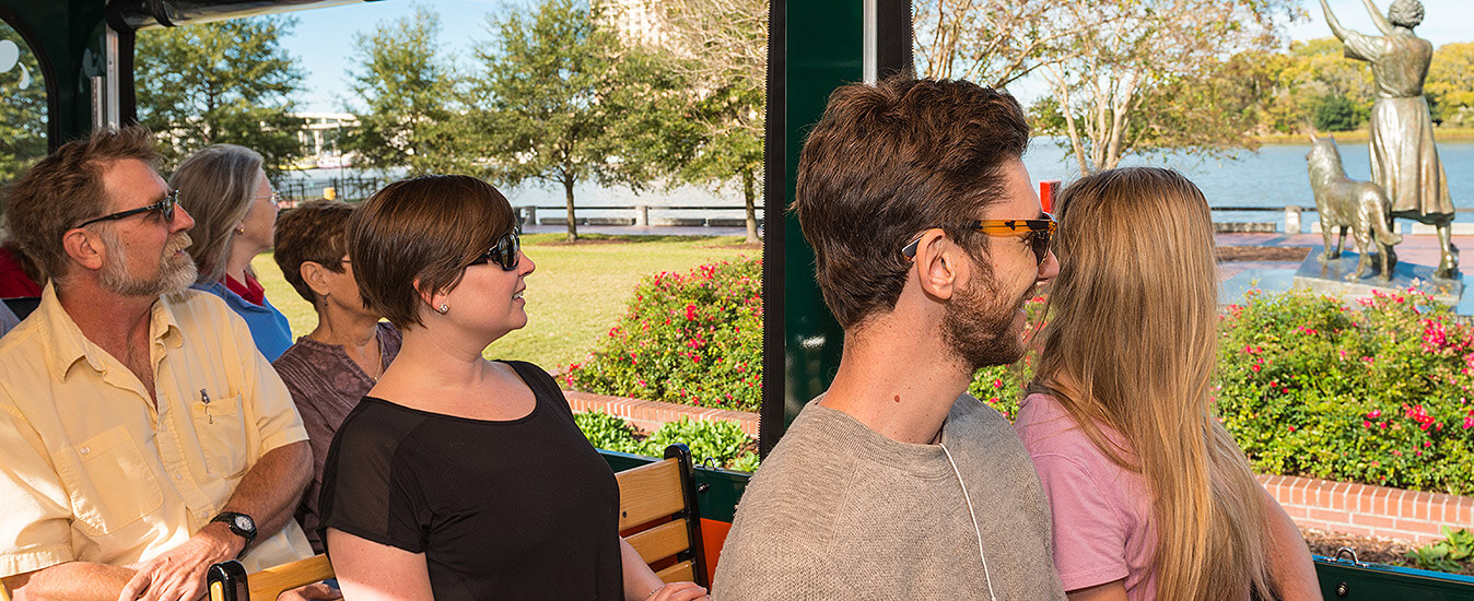 guests seated inside trolley looking out the window to views of the river and waving girl statue