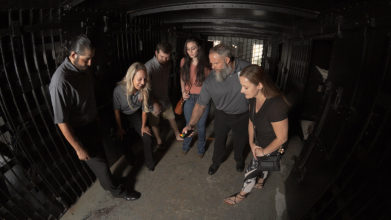 guests and tour guides standing around inside a cell at the St. Augustine Old Jail and they're all looking at an EMF meter held by one of the guides