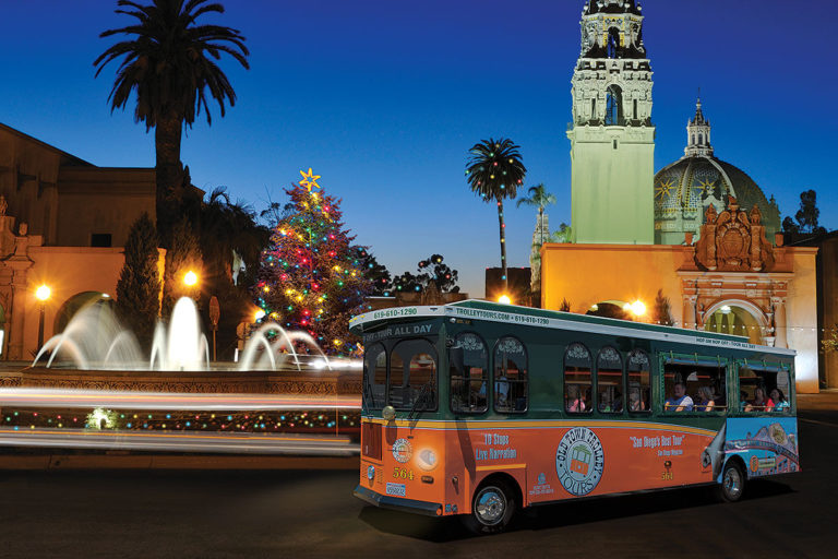 San Diego trolley driving at night past Balboa Park with a fountain, historical building and Christmas tree behind it